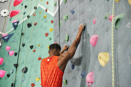 Foto de Vista trasera del hombre afroamericano en camisa naranja trepando por la pared de bouldering con cuerda de seguridad - Imagen libre de derechos