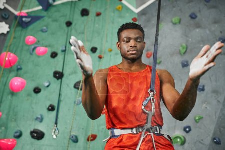 Photo for Young athletic african american man in orange shirt with safety rope using talc powder, bouldering - Royalty Free Image