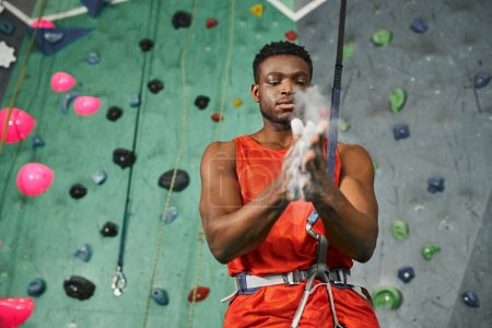 guapo atlético afroamericano hombre con camisa naranja usando tiza de gimnasio antes de ascender a la pared de roca