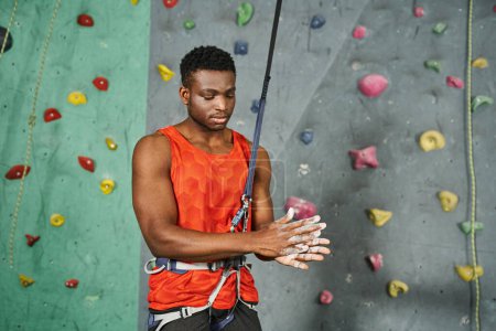Photo for Young athletic african american man in orange shirt with safety rope using talc powder, bouldering - Royalty Free Image