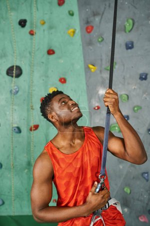Photo for Vertical shot of cheerful young african american man holding on safety rope, bouldering concept - Royalty Free Image
