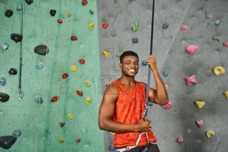 Photo for Cheerful sporty african american man in orange shirt smiling joyfully at camera, bouldering concept - Royalty Free Image