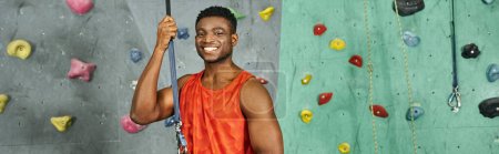 joyful athletic african american man in orange shirt smiling happily at camera, bouldering, banner