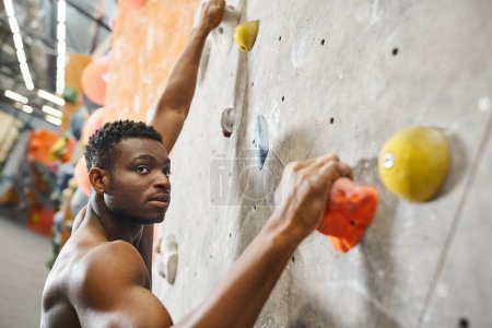 handsome fit african american man with his shirt off gripping on boulders while climbing up wall