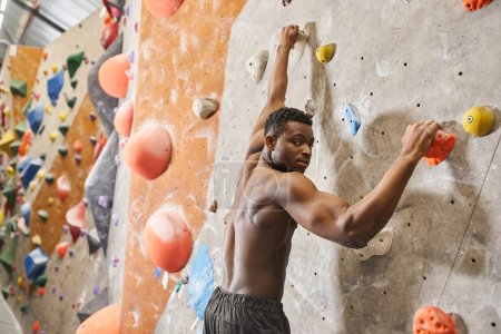 handsome shirtless african american man climbing up rock wall and looking at camera, bouldering