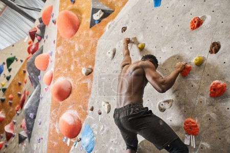 good looking fit african american man with his shirt off climbing up rock wall and looking down