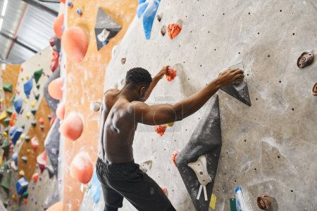sporty topless african american man posing on bouldering wall and gripping on rocks, looking down
