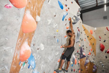 athletic shirtless african american man in black pants climbing up rock wall without safety rope