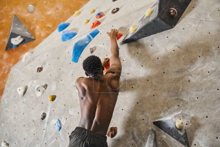sporty african american man with his shirt off reaching out boulder to grip, climbing up rock wall