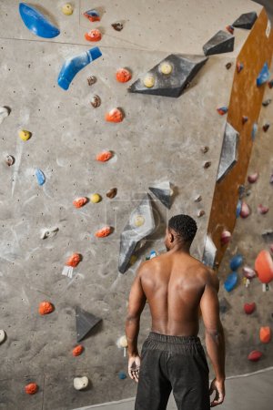 vertical shot of shirtless african american man in black pants posing next to bouldering wall