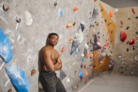 athletic african american man with shirt off posing leaning on bouldering wall, crossed arms