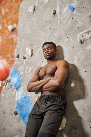 sporty good looking african american man posing topless next to climbing wall with arms crossed