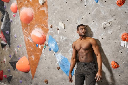 good looking shirtless african american man posing right next to climbing wall, smiling slightly