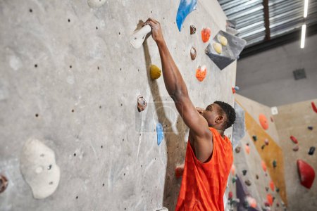 young african american man in orange shirt gripping on rocks while climbing up wall, bouldering