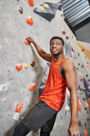 vertical shot of strong african american man smiling happily looking away while climbing up wall