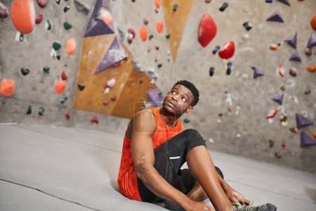 sportlicher junger afrikanisch-amerikanischer Mann in orangefarbenem Hemd sitzt neben Boulderwand und schaut weg