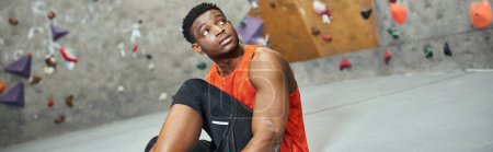 athletic african american man in orange shirt sitting next to rock wall and looking away, banner puzzle 675371518
