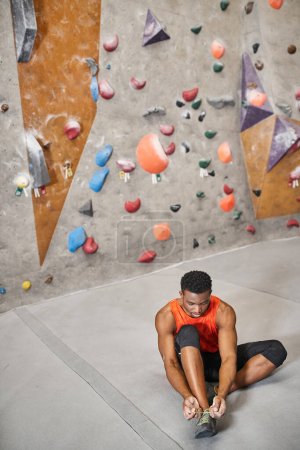 good looking african american man sitting on floor and tying his shoes next to climbing rock wall