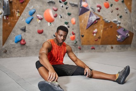 guapo joven afroamericano hombre de camisa naranja relajante en el suelo junto a la pared de bouldering