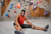 handsome young african american man in orange shirt relaxing on floor next to bouldering wall puzzle #675371546