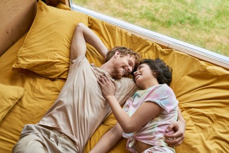 Ansicht von oben glücklicher Mann umarmt asiatische Freundin und liegt zusammen auf gelbem Bettzeug zu Hause, Paar