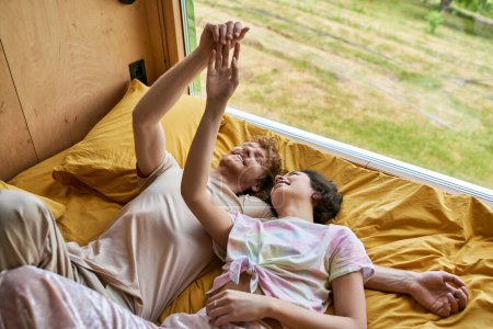 happy man and young asian woman touching hands and lying together on yellow bedding next to window