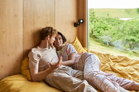 Photo for Redhead man talking to cheerful asian girlfriend while lying together in bed next to window - Royalty Free Image