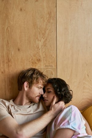 Téléchargez les photos : Couple interracial ayant moment tendre, rousse homme toucher la joue de femme asiatique et couché sur le lit - en image libre de droit