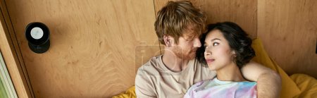 pelirroja hombre abrazando bastante asiático mujer y acostado en la cama cerca de ventana en casa de campo, pancarta