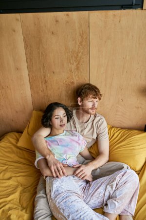 rousse homme câlin avec jolie asiatique femme sur confortable lit dans maison de campagne, tendres moments