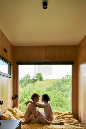 Foto de Escapada romántica, pareja multicultural mirándose mientras están sentados en la cama en una casa de campo - Imagen libre de derechos