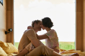 pretty asian woman kissing with redhead boyfriend and sitting on bed next to window with forest view Tank Top #675558532