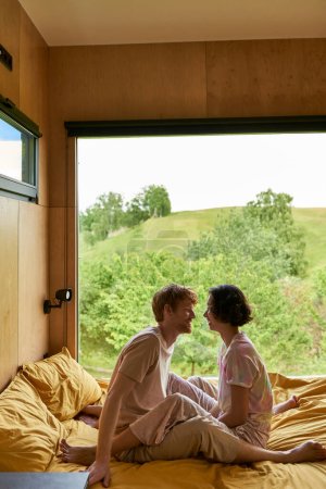 happy diverse couple looking at each other and sitting on bed in country house with forest view