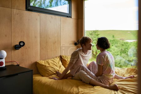 pareja multiétnica mirándose unos a otros y sentados en la cama en una casa de campo con vista al bosque