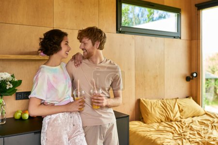 positive diverse couple standing in pajamas and holding glasses with orange juice in morning