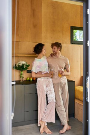 joyful barefoot couple standing in pajamas and holding glasses with orange juice in morning