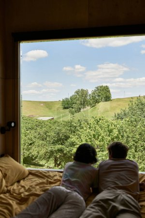 widok panoramiczny, widok z tyłu pary leżącej na łóżku i patrzącej na zielone drzewa na wzgórzu za oknem