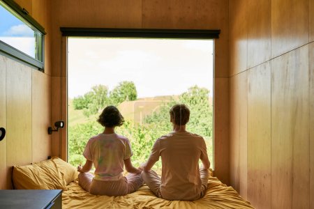 back view of couple meditating together and sitting on bed next to window with forest view