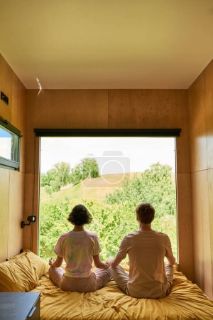 back view of couple sitting on bed and meditating together next to window with forest view