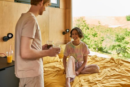 joyful asian woman sitting on bed and looking at redhead man with cups of coffee, happy morning time
