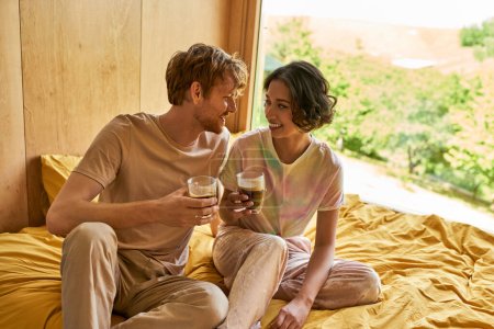 Photo for Happy interracial couple sitting together on bed and holding cups of morning coffee, weekend getaway - Royalty Free Image