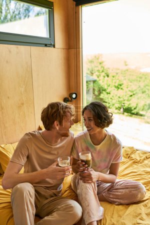 positive interracial couple sitting  on bed and holding cups of morning coffee, weekend vibes