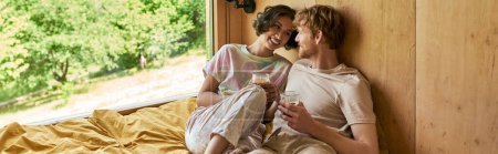 feliz pareja interracial sentado en la cama y la celebración de tazas de café de la mañana en la casa de campo, pancarta