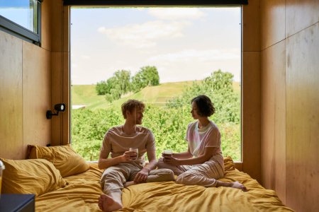 feliz pareja diversa sosteniendo tazas de café y sentado en la cama al lado de la ventana con hermosa vista