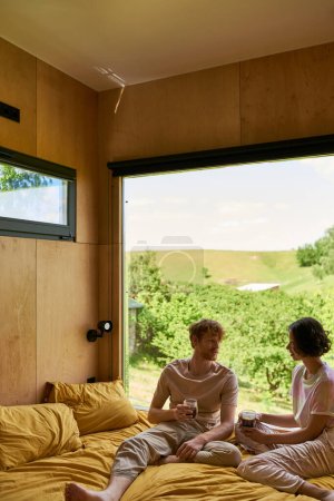 Téléchargez les photos : Couple multiculturel tenant des tasses de café et assis sur le lit à côté de la fenêtre avec une belle vue - en image libre de droit