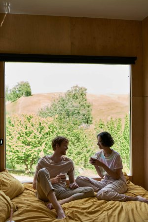 Téléchargez les photos : Heureux couple multiethnique tenant des tasses de café et assis sur le lit à côté de la fenêtre avec vue naturelle - en image libre de droit