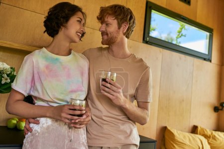 cheerful interracial couple standing in home wear and holding cups with morning coffee in kitchen