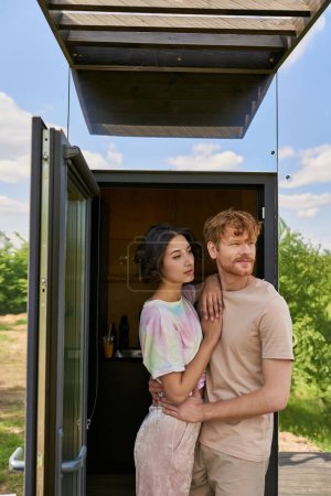 Téléchargez les photos : Heureux couple interracial debout à la porte d'entrée de la maison en verre moderne et regardant loin, l'air frais - en image libre de droit