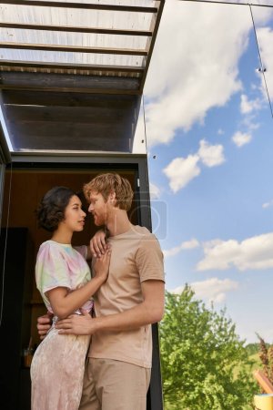 affectionate interracial couple standing at front door of glass house and looking at each other