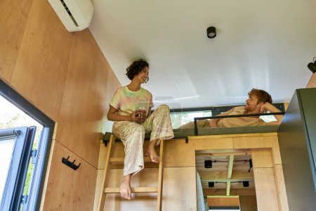 Photo for Happy asian woman holding cup of coffee and sitting at cozy bunk bed with redhead boyfriend - Royalty Free Image